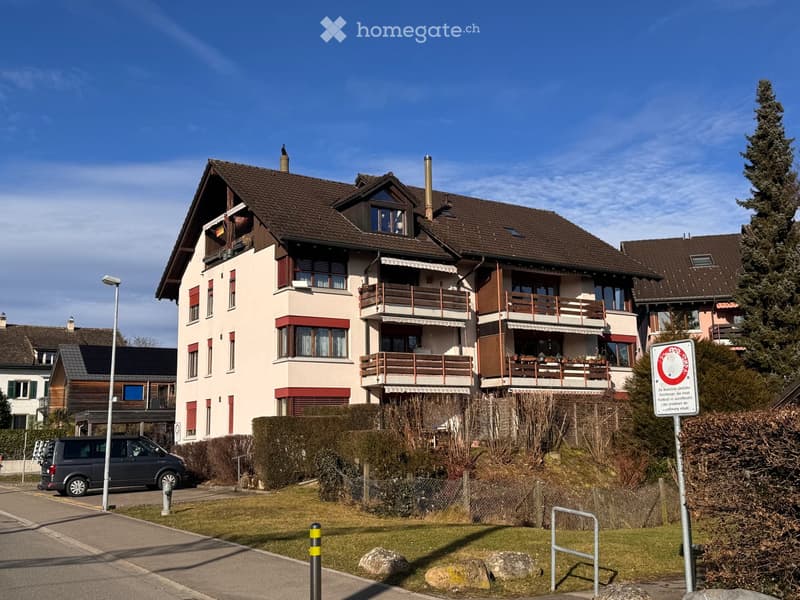 A two-story residential house with a brown tiled roof, multiple windows, balconies, and a well-maintained garden area.