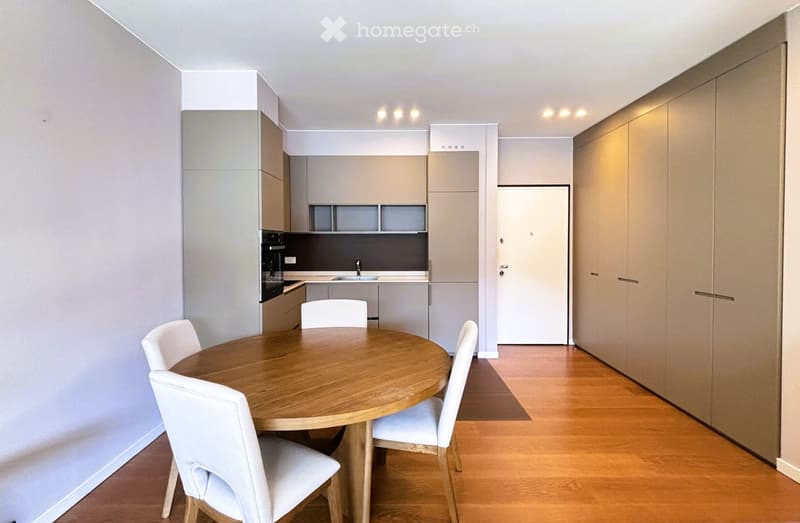 Dining area with a round wooden table and white chairs, modern kitchen with built-in appliances, and a hallway leading to a room.