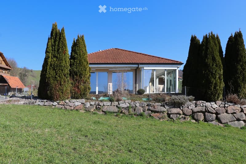 Detached house with a red roof, glass walls, surrounded by a stone wall and greenery