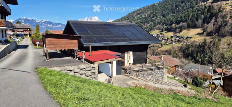Raised ground floor, solar panels on roof, wooden exterior, surrounded by greenery, mountain view