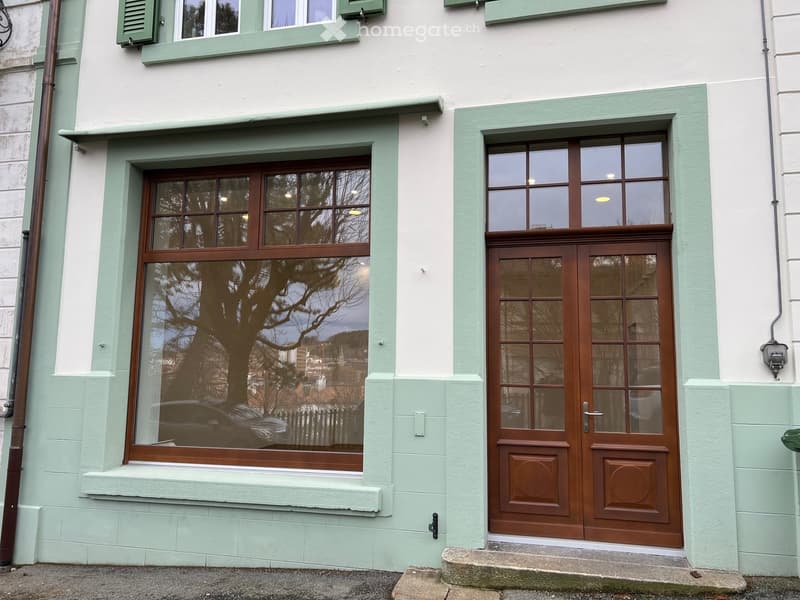 Facade of a house with green trim, large window with wooden frame, brown wooden door, steps leading to the entrance