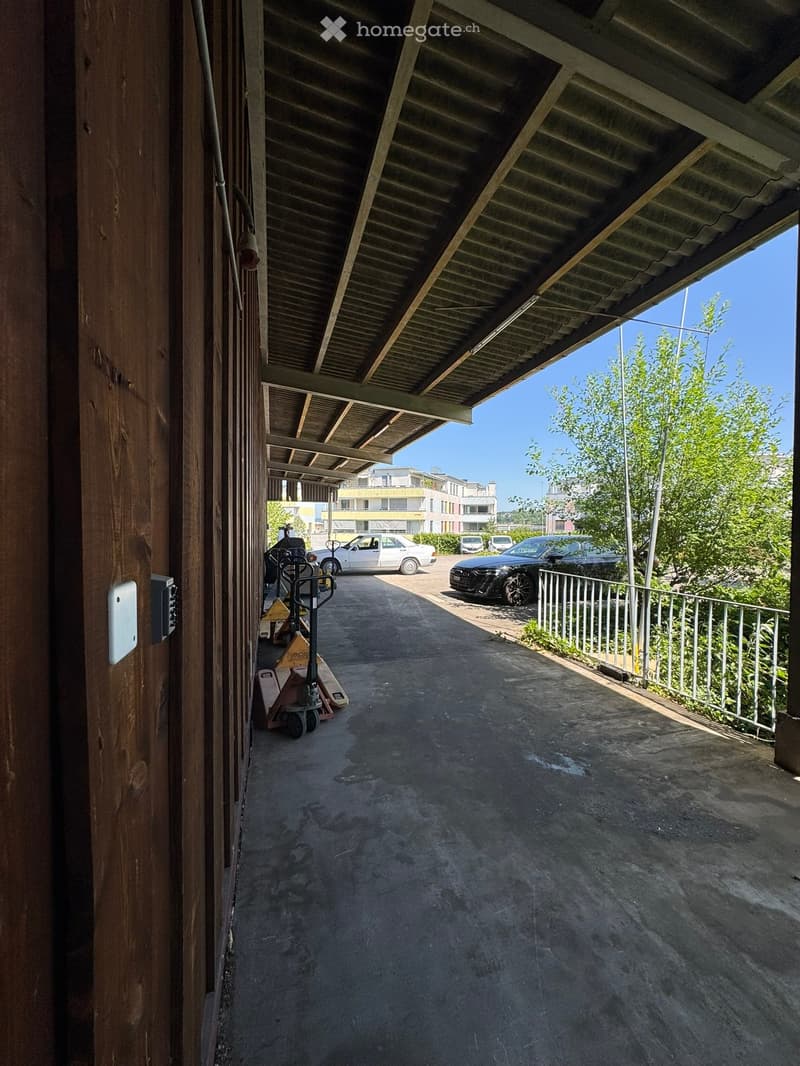 A covered outdoor corridor with a concrete floor, metal railing on one side, and several parked cars visible in the background.