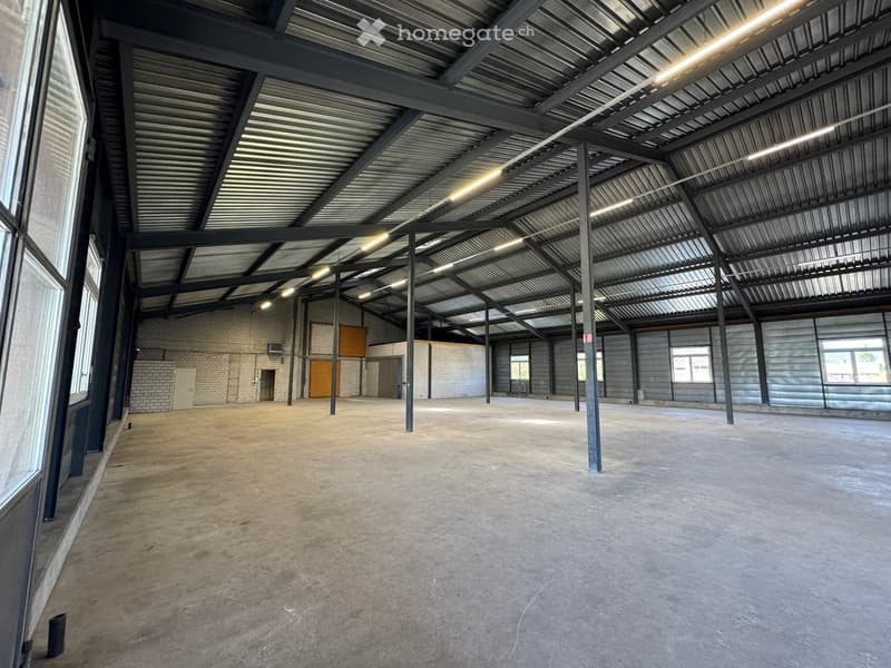 Large, empty industrial-style space with a concrete floor, metal ceiling, and exposed structural beams. The room has large windows on one side and a few doors leading to other areas.
