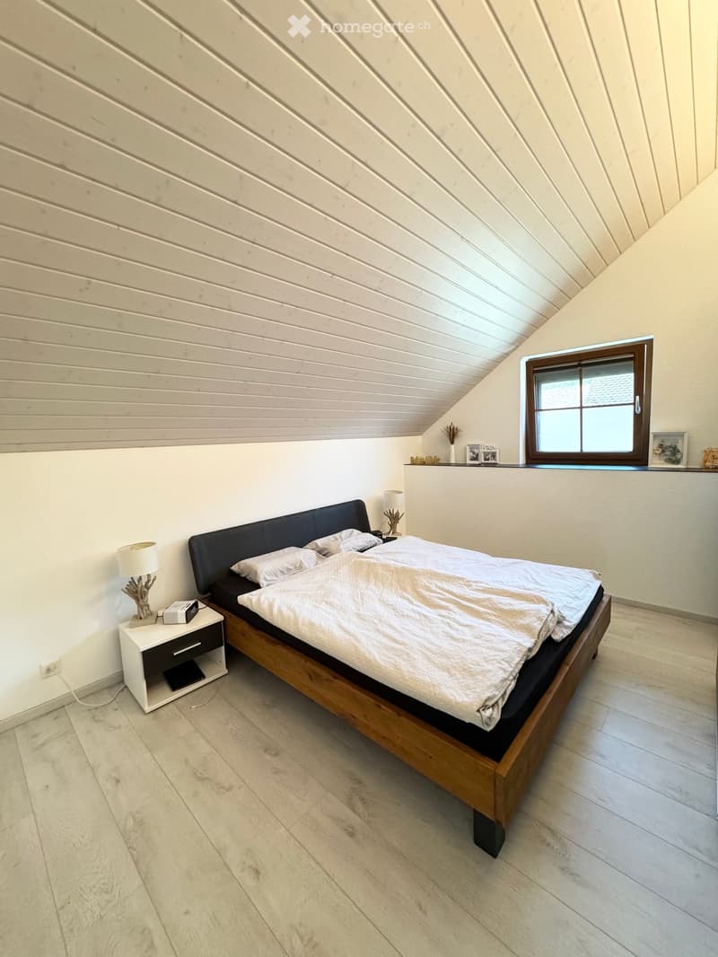 Bedroom with wooden bed, white bedding, two nightstands with lamps, white walls, wooden floor, window with white frame, white ceiling with wooden beams