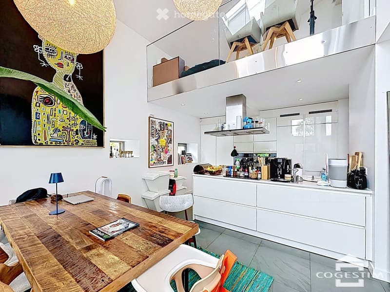 Open-plan living and dining area with a wooden dining table, chairs, a laptop, a lamp, and a modern white kitchen with appliances, cabinets, and a coffee machine.