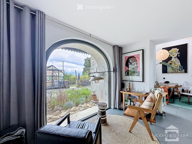 A modern living room with a large arched window offering a view of a garden, featuring a leather sofa, wooden chairs, a small table, a bar cart, and artwork on the walls.