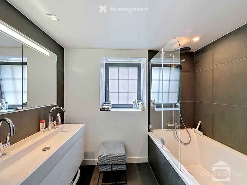 Modern bathroom with a double sink, bathtub with glass shower enclosure, and a window with towels and toiletries on the sill.