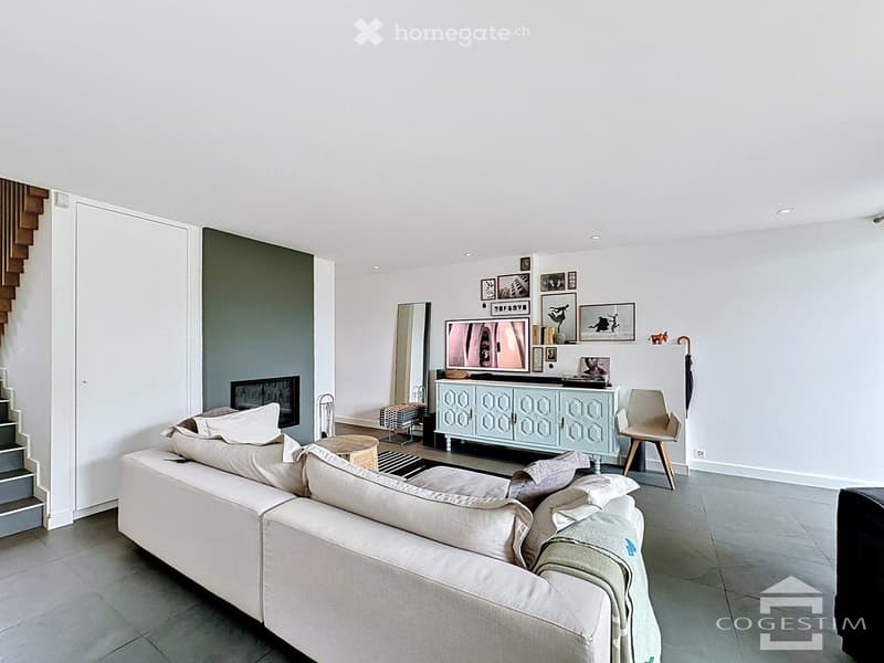 Modern living room with a large sectional sofa, coffee table, armchair, TV, cabinet, and mirror. Neutral tones with a grey accent wall and decorative artwork.