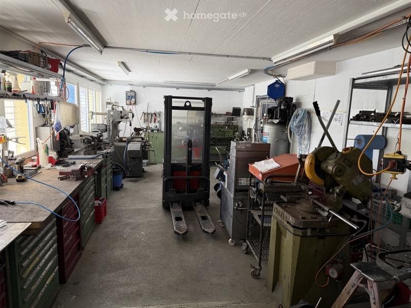 Industrial workshop with various machines, tools, and equipment. Forklift in the center, shelves with tools, and a table with a drill press.