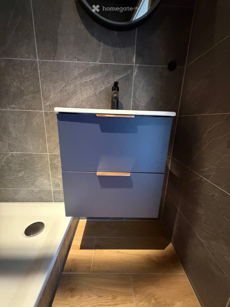Bathroom with blue cabinet, white countertop, black faucet, tiled walls, wooden floor, and a shower area.
