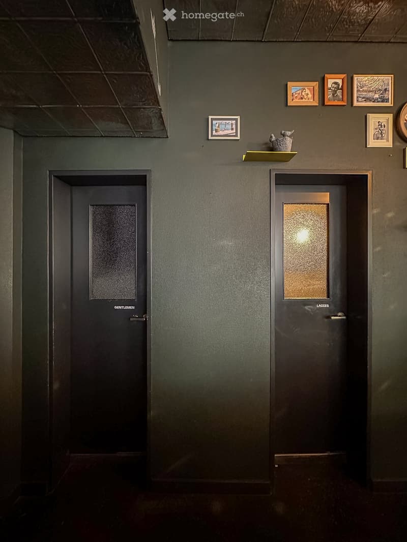 Two doors, one labeled 'Gentlemen' and the other 'Ladies', with glass panels, mounted on a green wall with picture frames and a shelf