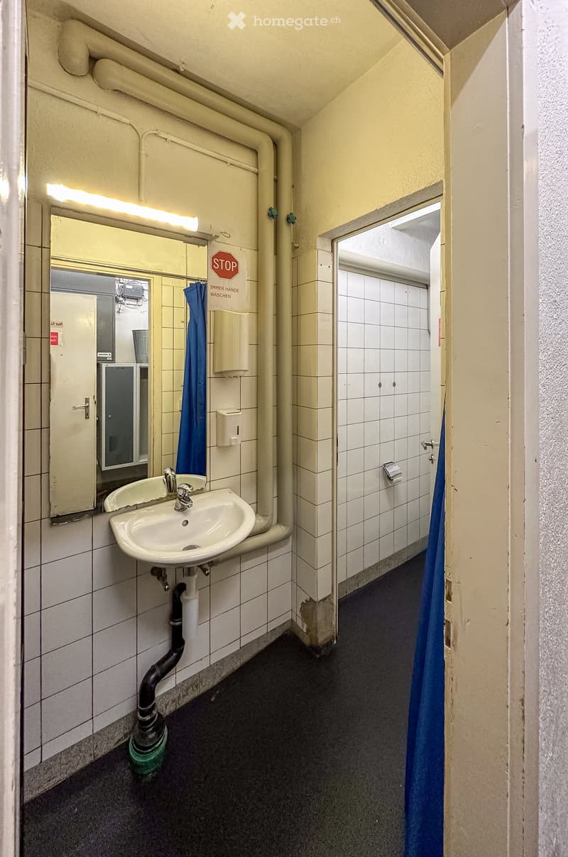 A small bathroom with a sink, mirror, and toilet. The walls are tiled, and there is a blue curtain for privacy.
