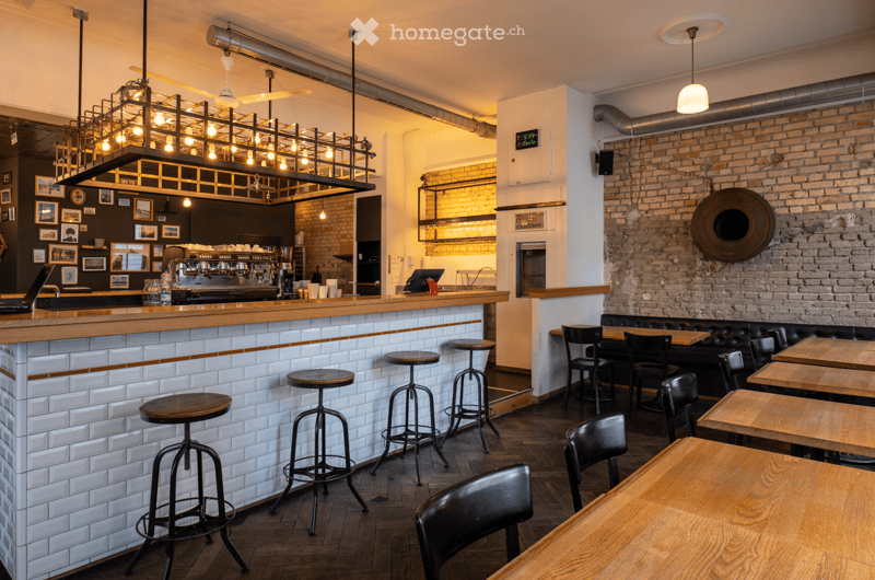 A coffee shop with a counter, stools, tables, chairs, brick wall, and a large chandelier.