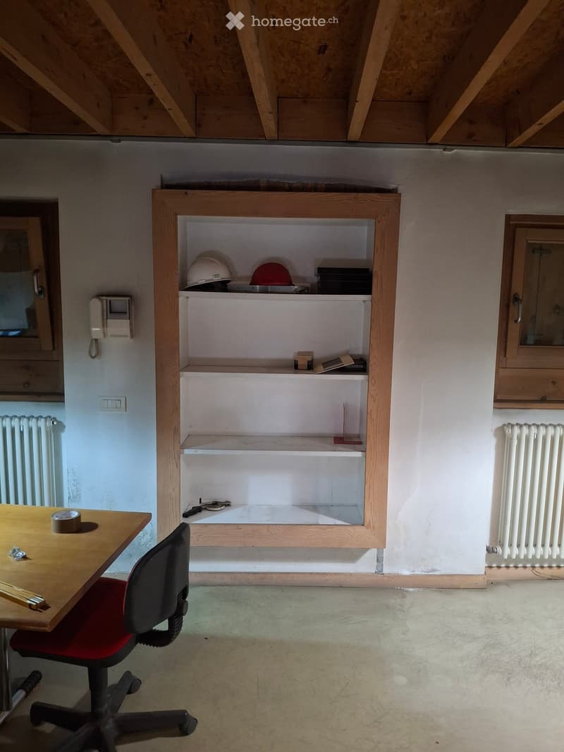 A small, unfurnished room with a wooden shelf containing helmets and tools, a table with a chair, and exposed wooden beams on the ceiling.