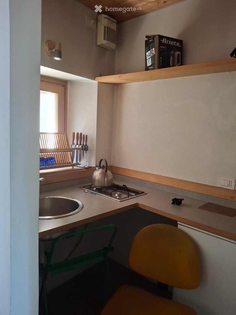 Compact kitchen with a sink, stove, teapot, knife rack, and a small table with two chairs. Wooden shelves and a wall-mounted lamp are visible.