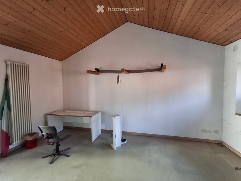 A small, unfurnished room with a wooden ceiling, a desk, a chair, a radiator, and a shelf. The floor is concrete, and the walls are white.