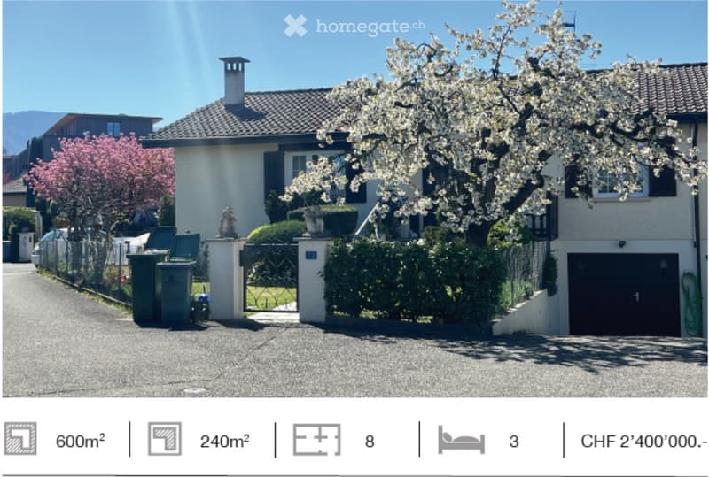 A two-story house with a tiled roof, white exterior walls, and black shutters. The house is surrounded by a garden with a variety of plants and trees, including a cherry blossom tree in full bloom. The driveway is paved with gravel, and there is a black g