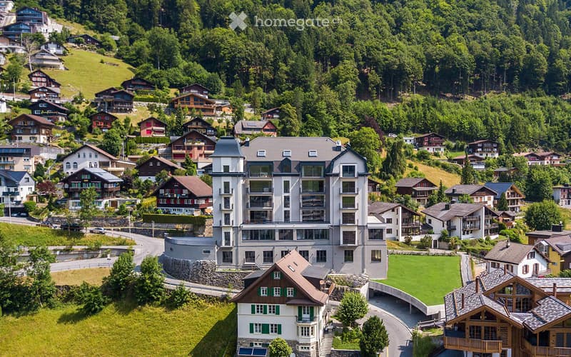 Aerial view of a modern apartment building surrounded by houses and trees, set on a hillside with a scenic valley below.