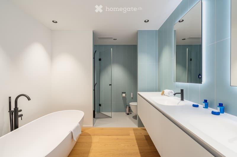 Modern bathroom with white bath, blue tiled walls, glass shower, white cabinets, sink, and black fixtures.