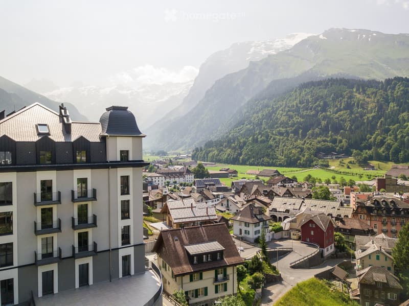 A modern multi-story building with balconies overlooking a picturesque village nestled in a valley surrounded by mountains and lush greenery.