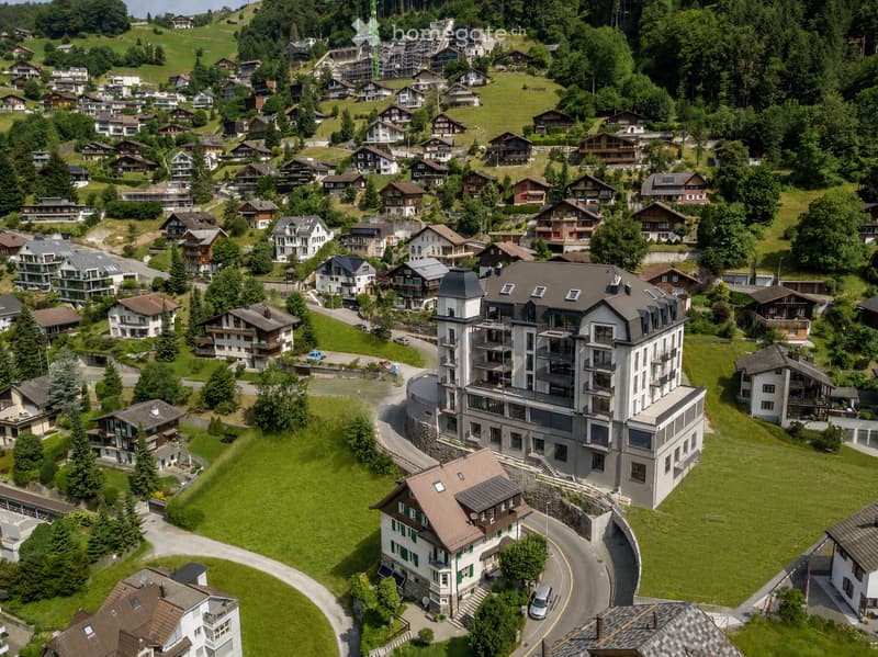 Aerial view of a residential area with multiple houses, green lawns, and trees, surrounded by hilly terrain and forested areas.