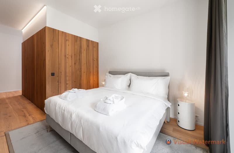 Contemporary bedroom with wooden wardrobe, white bed, white comforter, wooden floor, carpet, nightstand with lamp, light fixtures
