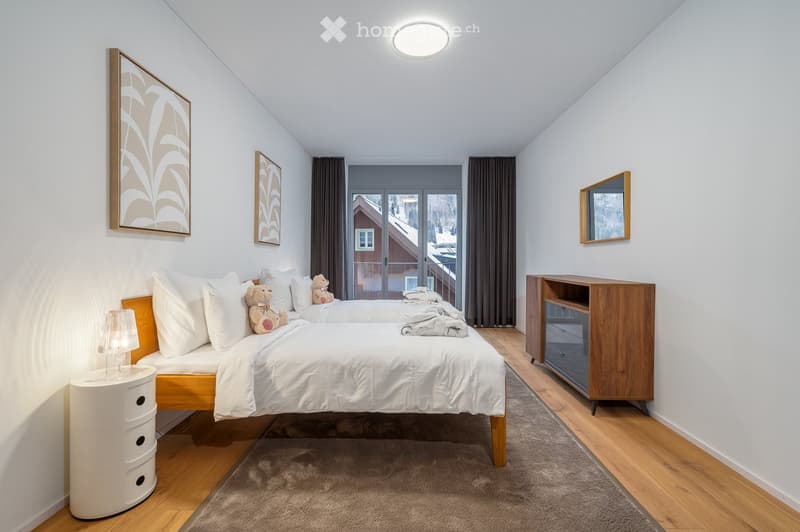 Bedroom with two beds, wooden floor, white walls, ceiling light, nightstand with lamp, wooden chest of drawers, small window, glass doors leading to balcony, artwork on the wall.