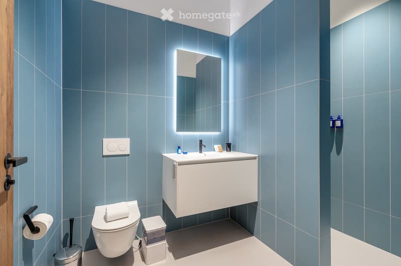 Blue tiled walls, white sink, modern mirror with lighting, white toilet, trash can, toilet paper holder, illuminated soap dispensers