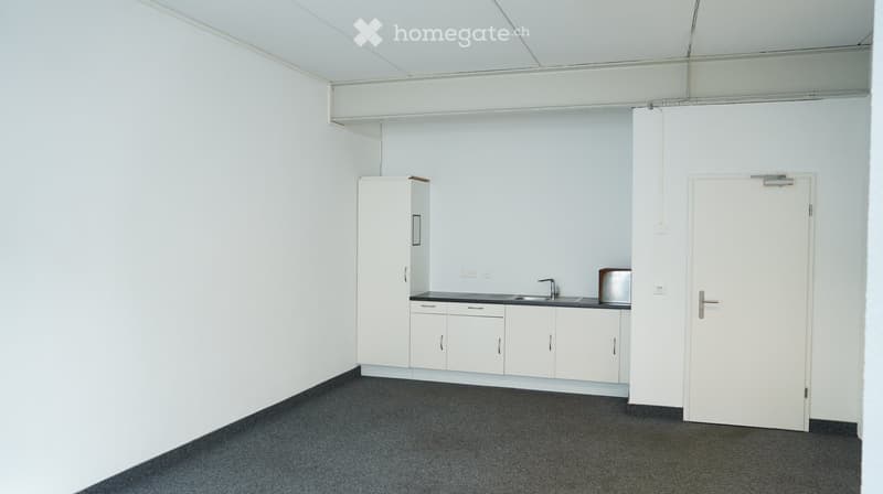An empty room with white walls, gray carpeted floor, and a kitchen area with cabinets, sink, and faucet.