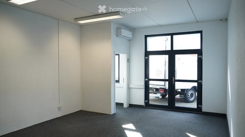 Empty room with white walls, gray carpet, and a glass door. There is a small window on the left wall and an air conditioning unit mounted on the wall above the door.