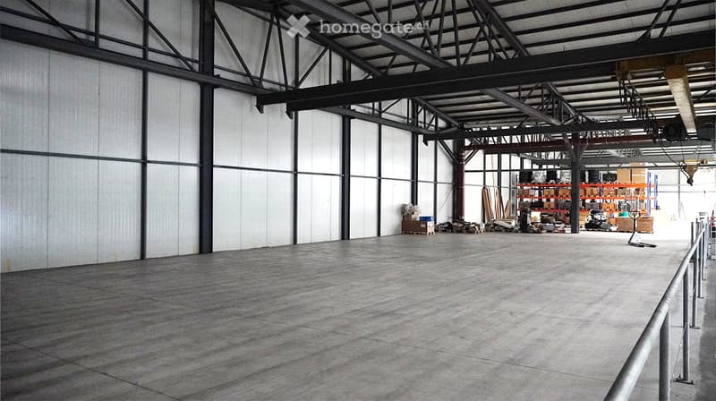 Empty warehouse with concrete floor, metal beams, and white walls. Storage boxes and equipment are present in the background.