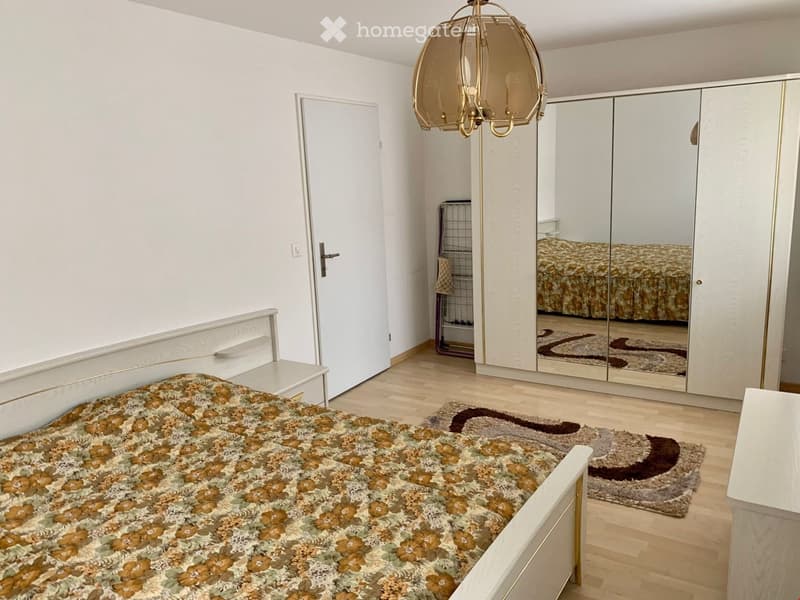 Spacious bedroom with a bed, nightstand, rug, wardrobe, lamp, mirror, and a rack. The room has wooden floors and white walls.