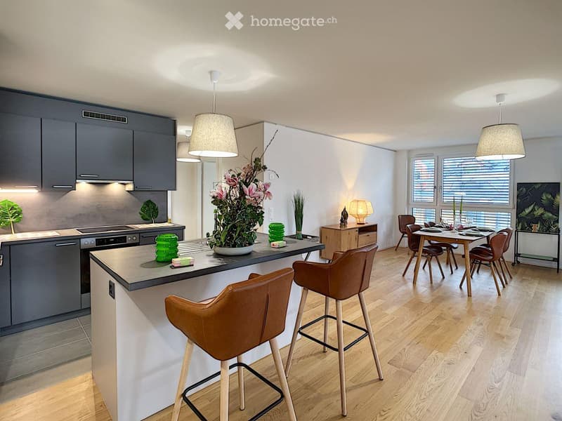 Modern kitchen with wooden floor, gray cabinets, and a large island with bar stools. The kitchen includes a stove, oven, and a sink. There is a dining area with a table and chairs. A plant and flowers are on the island, and a lamp is on the side table.