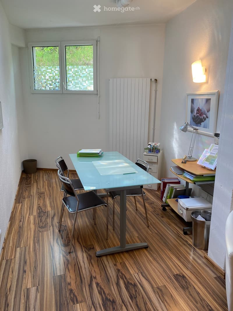 Furnished Room. A room with a desk, chairs, and a lamp. There are books on the desk and a trash bin nearby. A cabinet with drawers is against the wall, and a picture frame is mounted. A window with a view of grass is on the wall.