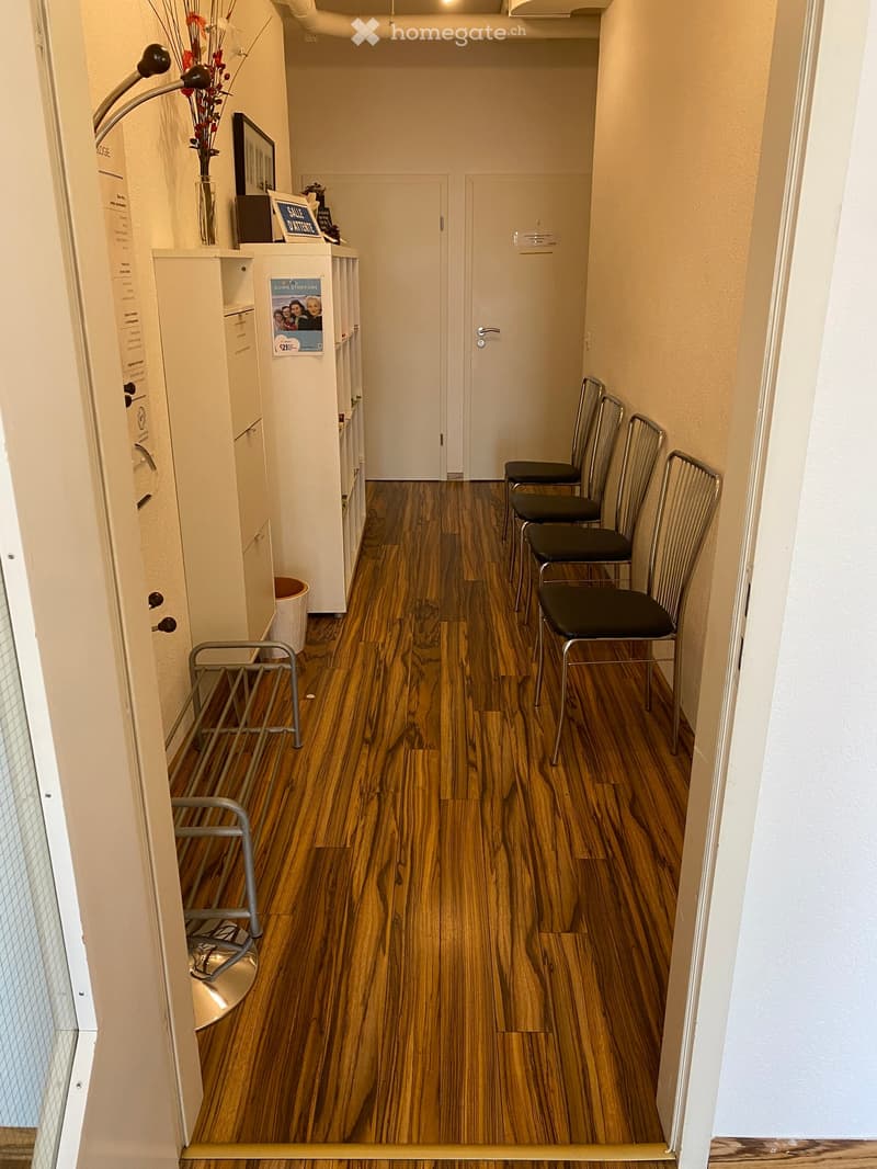 Other. {"visible_features":[],""type":"Corridor / Hallway",""objects":["chairs","cabinet","trash bin","vase","flowers","poster","picture frame","book","sign"],""description":"A hallway with wooden floors, a row of chairs, a cabinet with a vase of flowers,