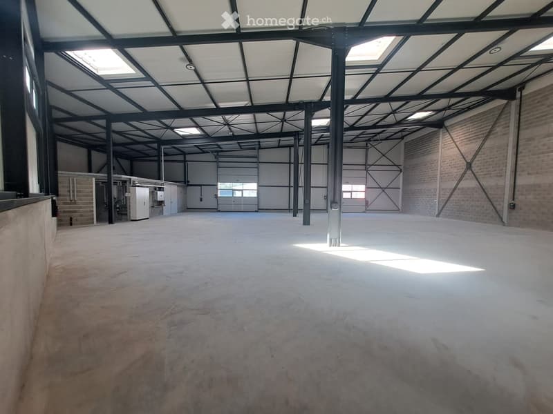 Large, empty industrial-style space with concrete floor, exposed metal beams, and large windows allowing natural light. Minimal furnishings, including a small utility area with cabinets and equipment.