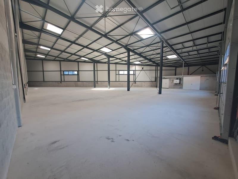 Large, empty industrial-style space with a high, metal-framed ceiling and concrete floor. Minimal lighting and no furniture or equipment.