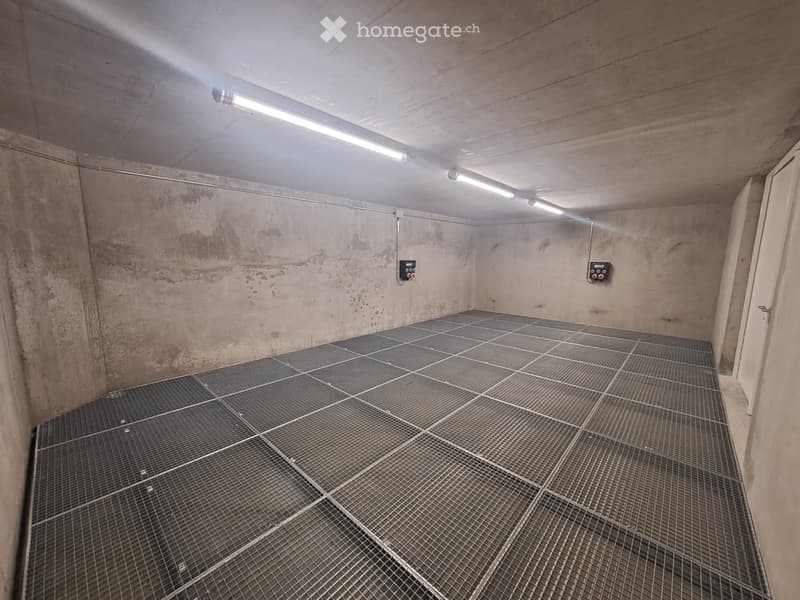 Empty concrete-walled room with a metal grated floor and fluorescent lighting, showing no furniture or equipment.