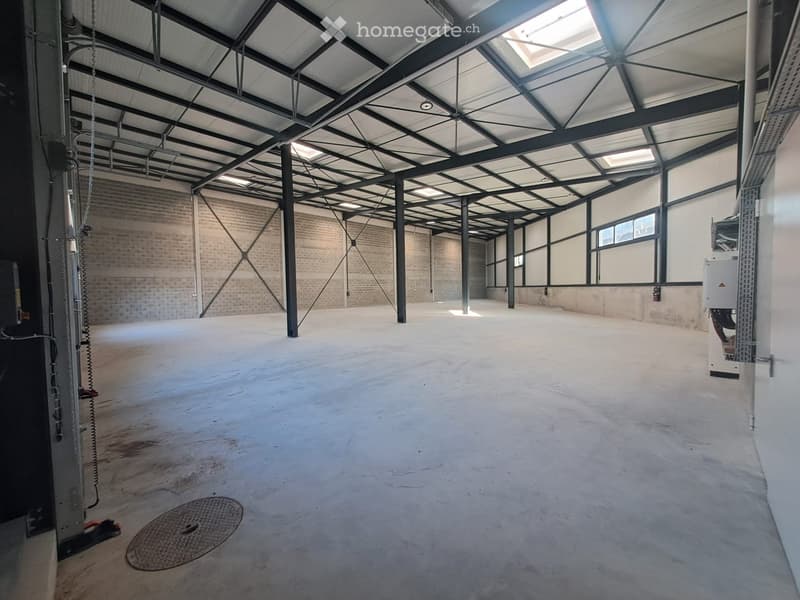 Empty industrial-style room with exposed metal beams, concrete floor, and large windows, featuring a manhole cover and air conditioning unit.