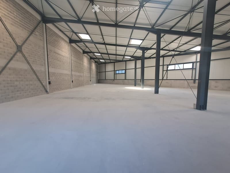 Empty industrial-style room with exposed brick walls, metal beams, and a concrete floor. The space is well-lit with natural light from large windows and a skylight.