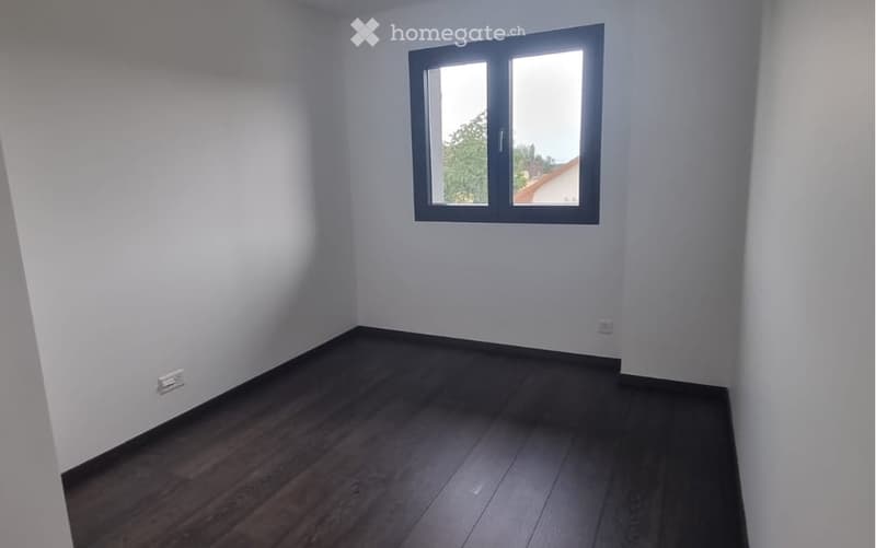 Empty room with white walls, dark wooden flooring, and a single window with a black frame.