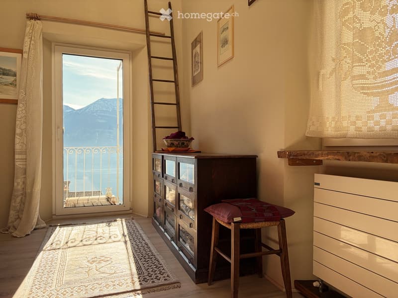 A room with wooden floor, a glass door leading to a balcony, a wooden ladder, a wooden cabinet with glass doors, a stool with a pillow, curtains, picture frames on the wall, and a radiator