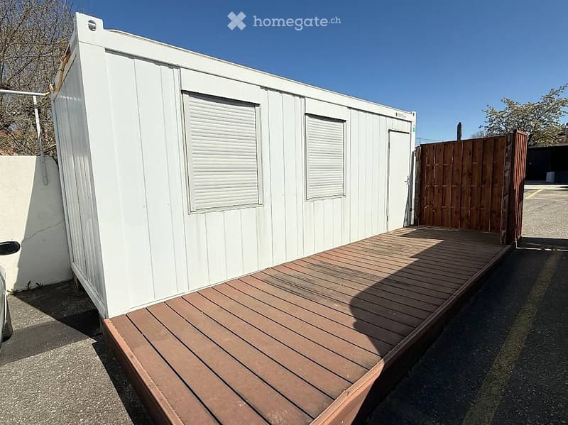 Building. A white, rectangular shipping container with a wooden deck attached to its side, featuring two windows with blinds and a door.