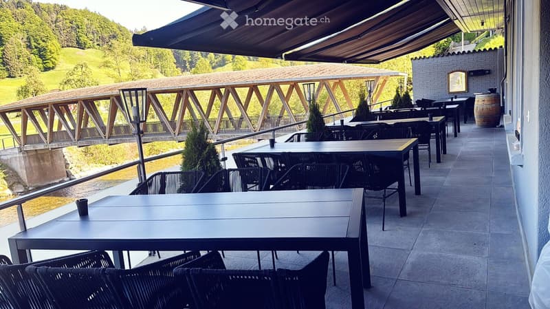 Outdoor terrace with tables and chairs, overlooking a river and wooden bridge, with a covered awning and scenic view.