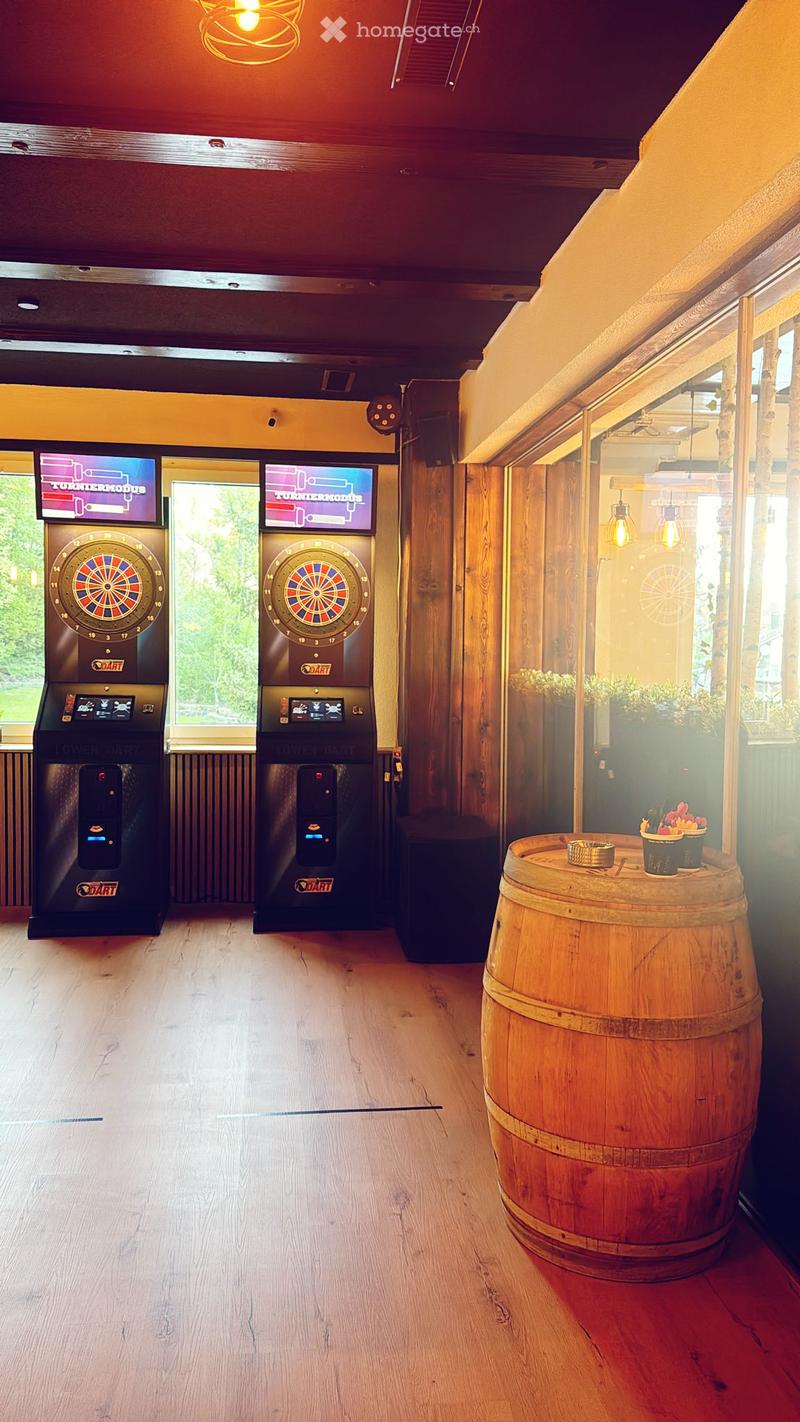 A modern, well-lit room with wooden flooring, two dartboards with screens, a large wooden barrel as a table, and a small potted plant on the barrel.