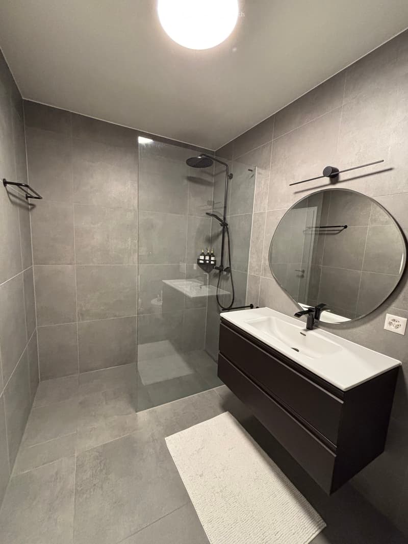 Bathroom. Modern bathroom with a white sink, dark vanity, large round mirror, glass-enclosed shower, and tiled walls and floor.
