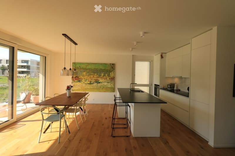 Kitchen. Modern kitchen with light wood flooring, white cabinets, a central island, and a dining area with a wooden table and chairs. Large windows provide natural light, and pendant lights hang above the island.