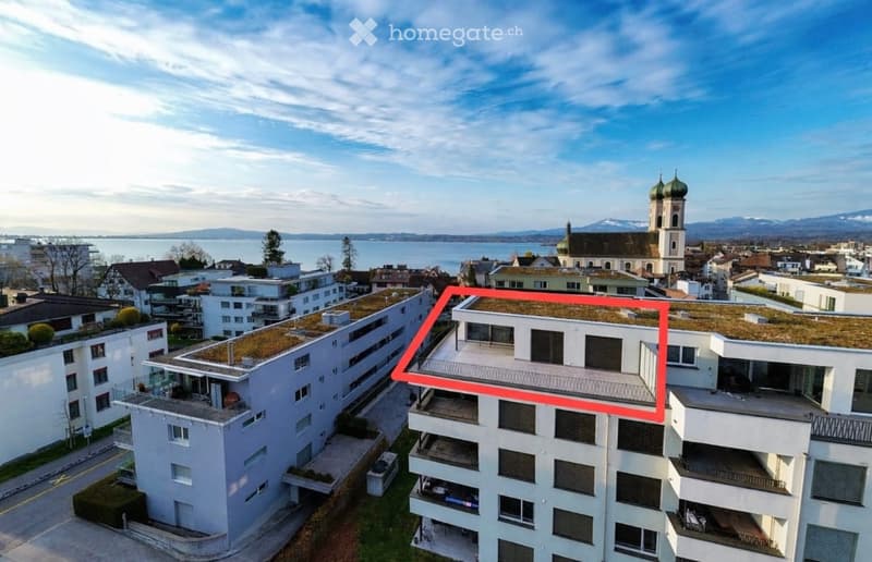 Building. Aerial view of a modern residential complex with multiple buildings, green roofs, balconies, and a scenic backdrop of a lake and distant mountains.