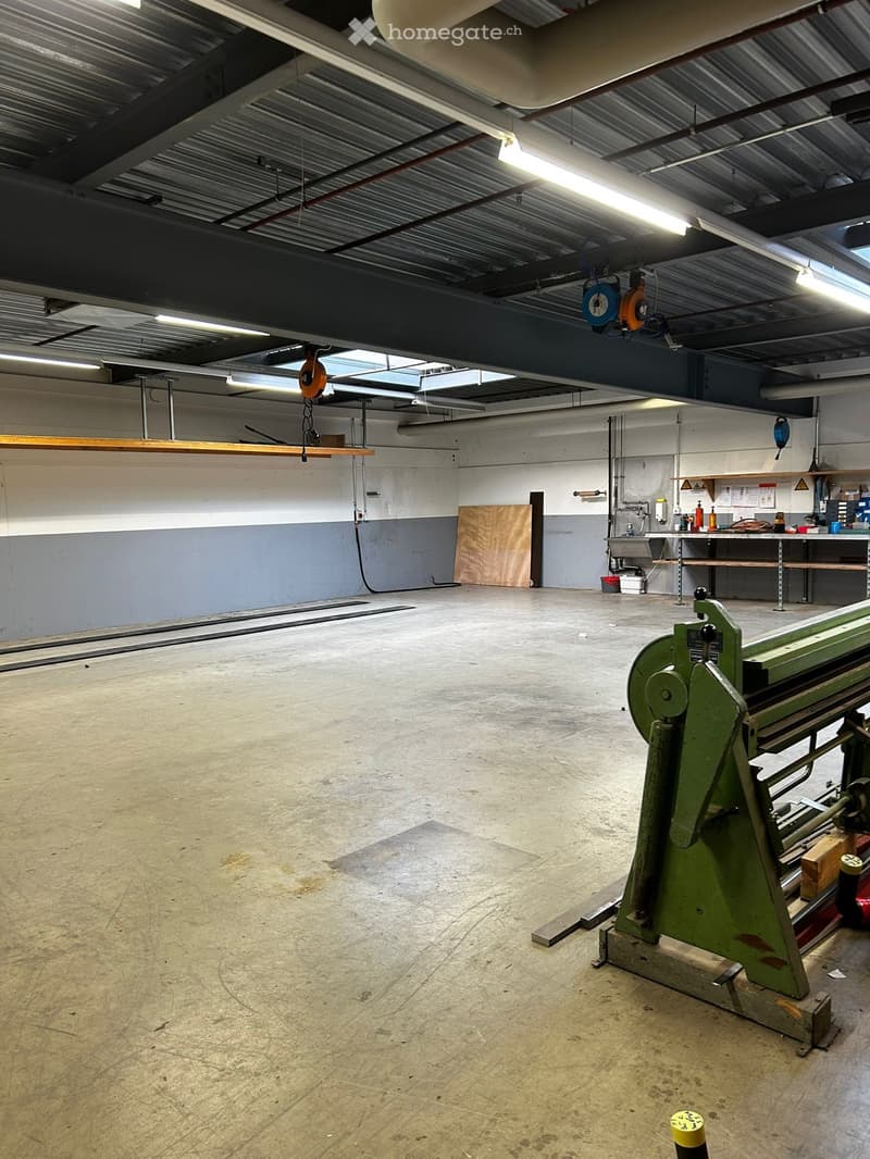 Unfurnished Room. Large, empty workshop or garage space with concrete floors, exposed metal beams, and industrial lighting. Features a workbench with tools, a large green industrial machine, and a wooden plank leaning against the wall.
