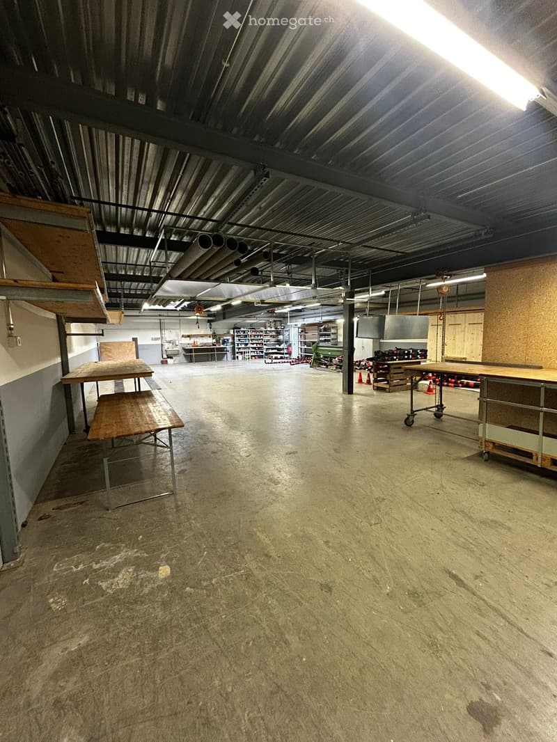 Furnished Room. Large industrial-style room with exposed metal ceiling, concrete floor, and minimal furnishings including a wooden table, bench, and shelves.